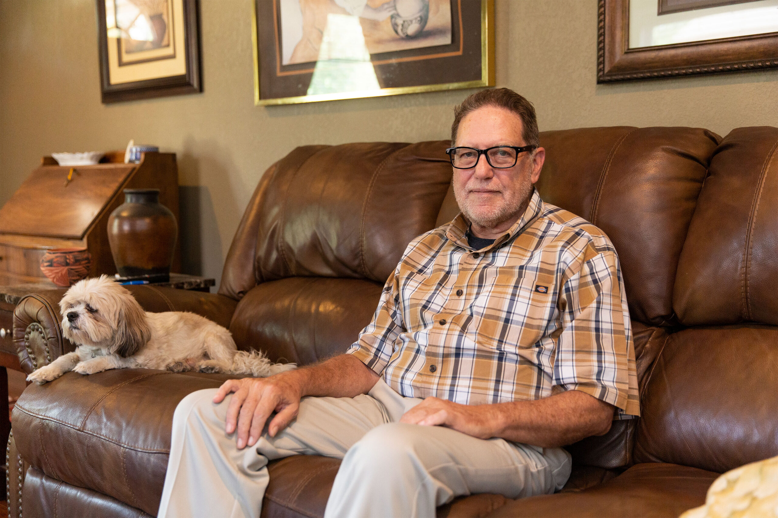 A man in a checkered shirt sits on a brown couch in a living room. A small white and brown dog sits beside him.